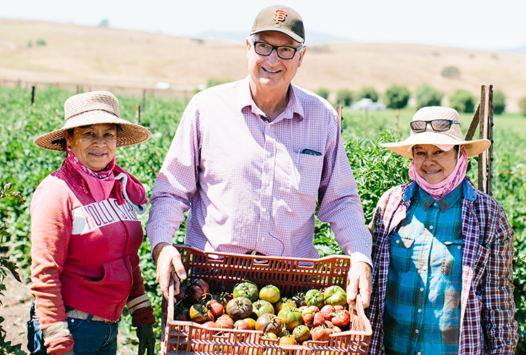 3 farmers holding a basket of crops in front of a field