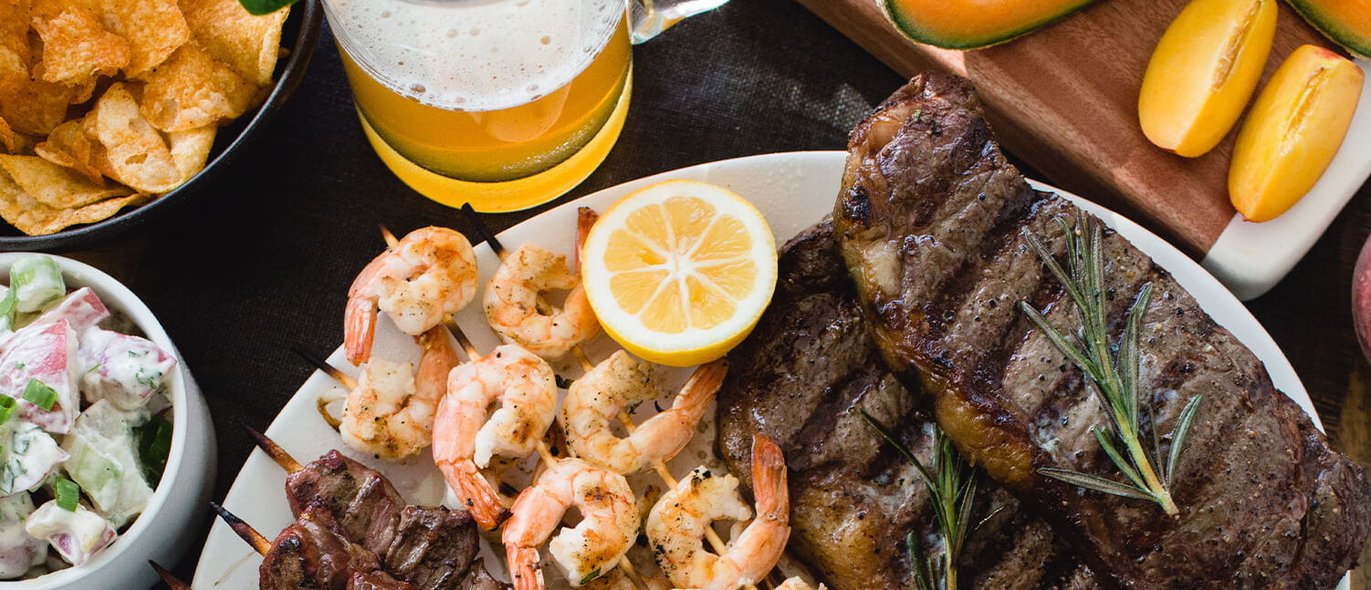 a plate with skewers of shrimp and meat, steaks, herbs and lemon with sliced fruit, deli sides and beer in the background.